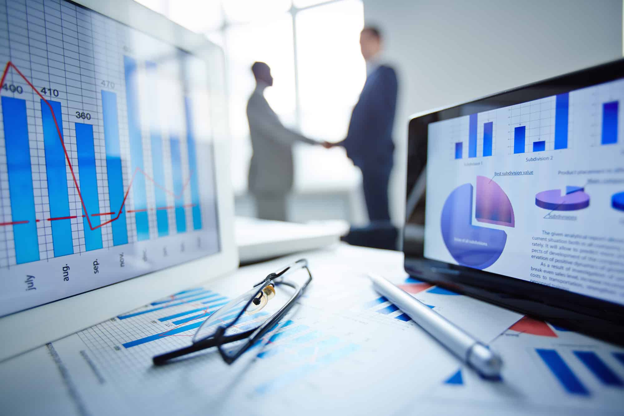 Image of eyeglasses, pen, two touchpads and financial documents at workplace with businessmen handshaking on background